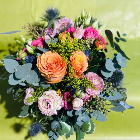 Floraler Überraschungsstrauß mit orangefarbenen Rosen, pinken Blüten, Edeldisteln, Eukalyptus und zartem Beiwerk vor grünem Hintergrund