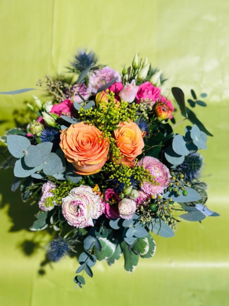 Floraler Überraschungsstrauß mit orangefarbenen Rosen, pinken Blüten, Edeldisteln, Eukalyptus und zartem Beiwerk vor grünem Hintergrund